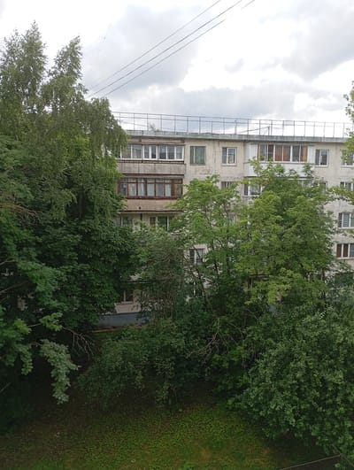 The image shows a multi-story apartment building partially obscured by dense green trees and foliage. The building appears to be made of concrete and has numerous windows and balconies. Some windows are open, revealing blinds or curtains. A metal railing is visible on the roof of the building. Overhead, several power lines stretch across the cloudy sky. The ground in front of the building is covered with grass and scattered leaves. The scene is consistent with an urban environment in Gorki-2, Russia, likely during the summer or late spring due to the lush vegetation. There are no people visible in the image.