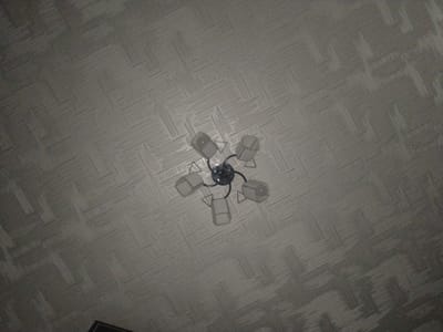 The image shows a chandelier with five lights attached to a textured ceiling. The chandelier has a circular base and arms that extend outwards, each holding a light fixture with a white, frosted glass shade. The ceiling has a repeating woven pattern in a light gray color. The image is taken from below, looking directly up at the ceiling. The location of the scene is in Zarafshan City, Uzbekistan.