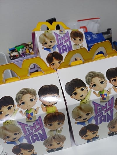 This indoor image, located in 서대문구, South Korea, displays three McDonald's Happy Meal boxes. Two white cardboard boxes dominate the foreground, positioned side-by-side with the left box slightly angled forward. Each box features the iconic yellow arch handles and is adorned with colorful illustrations of multiple chibi-style animated characters from the "TinyTAN" franchise, along with the prominent "TinyTAN" logo. These characters are depicted in various poses. A third, partially visible McDonald's box is situated further back, behind the two foreground boxes. To the left in the midground, a cluster of small, colorful figurines, including characters resembling Batman and the Joker, are arranged on a purple shelf or surface. The background consists of a plain, light-colored wall, with a white plastic bag, a dark rectangular object, and a blue packet with Korean text partially visible. The scene is illuminated by bright, even lighting, which produces a reflective glare on the glossy surfaces of the boxes.