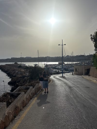 Two individuals, one appearing to be a woman with dark hair wearing a dark t-shirt and shorts, and another person with lighter hair wearing a white top and dark trousers, are walking on a paved road. The person with dark hair is holding a smartphone and appears to be taking a photograph or video. They are walking away from the camera, towards a marina filled with numerous boats, including sailboats and smaller motorboats. The marina is situated on the coast, with a breakwater made of large stones extending into the water on the left side of the image. A low stone wall with planters runs alongside the road on the left. In the background, a town with buildings and vegetation is visible on a hill overlooking the sea. The sun is high in the sky, creating a bright glare and casting long shadows on the road. The overall scene suggests a daytime view in a coastal town, possibly in the Mediterranean. The location is Urbanización Cabo Roig, Spain.