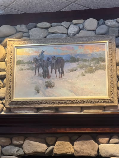 This is a medium shot of a painting of two cowboys on horseback in a snowy landscape. The painting is framed in a wooden frame with a distressed finish and is mounted on a stone wall. The cowboys are wearing hats and chaps, and one is holding a rifle. The landscape is barren and covered in snow, with a few bushes and trees in the background. The sky is a warm orange and pink, suggesting a sunset. The painting is signed in the bottom right corner, but the signature is illegible. The stone wall is made up of large, rounded river stones of various colors, including gray, tan, and white. Above the stone wall is a dark brown wooden mantelpiece. The ceiling above the stone wall is a textured drop ceiling. The overall impression is one of a rustic and Western-themed setting, possibly in a restaurant or home. The location is identified as the city of Jackson, United States of America.
