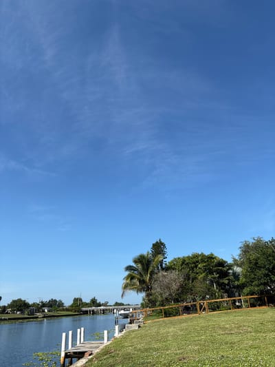 The image depicts a serene canal scene in North Port, Florida. A calm, blue canal runs horizontally across the lower left of the frame, bordered by lush green grass on the right. A wooden dock with white pilings extends into the water from the foreground. Across the canal, a residential area with houses and trees is visible, with a bridge spanning the waterway in the mid-ground. A white boat is docked on the right side of the canal. The sky is a clear, bright blue with a few wispy white clouds. The overall impression is one of peaceful, tropical living.