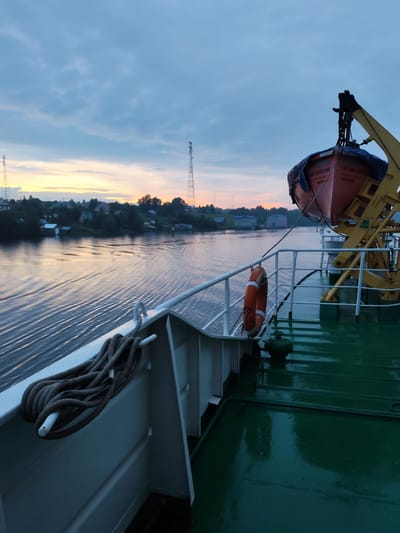 The image captures a twilight view from the deck of a vessel navigating a calm river, likely in the vicinity of Ivanov Bor, Russia, given the presence of characteristic infrastructure. In the foreground, a thick, coiled rope rests on a white railing, a common feature on maritime vessels. To the right, a bright orange life preserver hangs on the railing, ready for use. Further back, a red lifeboat is secured to a yellow davit, a structure designed for lowering boats.
The river stretches into the distance, its surface reflecting the soft hues of the sunset sky, transitioning from warm oranges and yellows near the horizon to cooler blues and grays overhead. The water exhibits gentle ripples, indicating the vessel's movement. Along the riverbank, a settlement is visible, with houses and trees dotting the landscape. Two prominent telecommunication towers stand tall against the sky, one closer and more detailed than the other. In the middle ground, a few utilitarian buildings with blue roofs suggest industrial or commercial activity. The overall atmosphere is serene and tranquil, characteristic of a peaceful evening on the water.