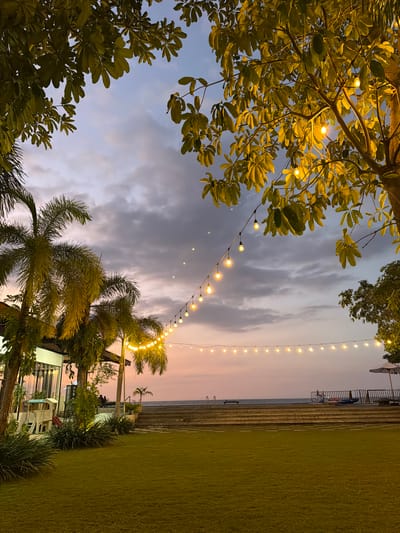 This image captures an outdoor scene at dusk, likely a resort or recreational area, near Pasir Putih, Indonesia. The foreground is dominated by lush green grass on the right and the dark silhouettes of trees and palm fronds on the left and bottom. A string of warm, glowing lights hangs across the frame, illuminating the foliage and adding a festive ambiance.
In the background, a tiered structure resembling a staircase or seating area leads down towards the water. The sky is a soft gradient of twilight colors, with hues of pale pink and purple blending into the dusky blue. Wispy clouds are scattered across the sky. A calm body of water, likely the sea, is visible in the distance beyond the tiered area. A few people are seated in the distance, enjoying the evening.
The overall mood is serene and tranquil, with the gentle glow of the lights and the soft colors of the sky suggesting a peaceful end to the day. The presence of palm trees and the overall tropical feel of the vegetation strongly hint at a coastal location in a warm climate. There is no visible text in the image.