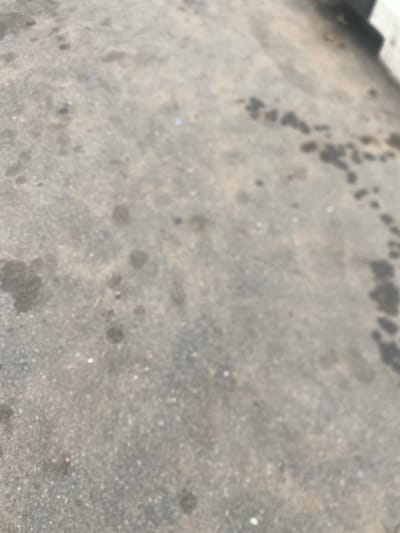 The image depicts a blurred asphalt surface, likely a road or parking lot. There are several dark, irregular stains scattered across the surface, which appear to be oil or grease. The lighting suggests it is daytime. There is no indication of the location in Gulum, Nigeria, as the image lacks any distinctive landmarks or identifying features. The scene is devoid of people or any specific objects that would provide further context.