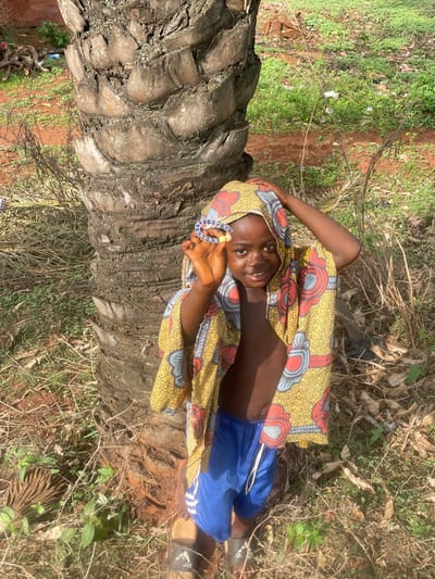 A young person is lying on a fallen palm tree trunk in an outdoor environment. The person is wearing blue shorts and a head wrap and robe featuring a yellow, red, and blue patterned fabric. They are holding a beaded bracelet in their raised hand, smiling, and looking at the camera. The palm tree trunk is brown and textured. The ground is covered with grass, leaves, and other vegetation. The location is Ogbe, Nigeria.
