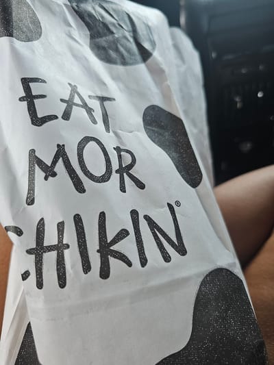 A close-up, slightly angled shot of a white paper Chick-fil-A bag with black speckled cow spots and black text. The text on the bag reads "EAT MOR CHIKIN". The bag is held by a person whose arm and leg are visible on the right side of the frame. The background is dark and out of focus, suggesting the image was taken inside a vehicle. The location is not specified in the image, but if it were in Homestead, United States of America, it would be a common sight at a local fast-food restaurant.