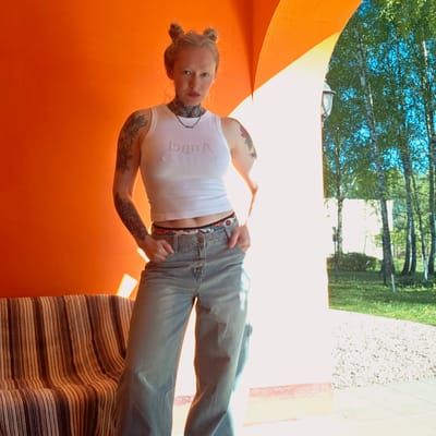 A woman with blonde hair styled in two buns is standing indoors in front of a bright orange wall. She is wearing a white tank top with text, light wash jeans, and a patterned belt. Her left arm and chest have tattoos. The location appears to be in Тэльмінскі сельскі Савет, Belarus, as evidenced by environmental features visible through a partially open doorway or window. These include trees, green vegetation, and a light-colored path or driveway. A striped rug or blanket is partially visible at the bottom right of the image.