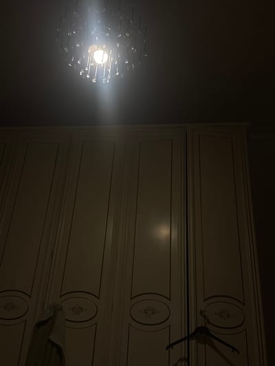 The image shows a poorly lit interior space in Perugia, Italy. A chandelier with a bright, single bulb is the primary light source, casting a strong glare. To the right, a large, white dresser with multiple drawers is visible. The drawers have decorative oval designs with floral motifs. A clothes hanger is partially visible, hanging from the top drawer. The background is dark, suggesting a nighttime scene or a room with minimal lighting. A folded blanket or piece of clothing is partially visible on the dresser.