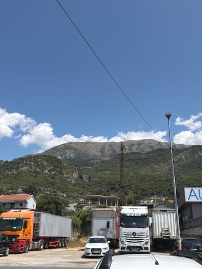 The image depicts a street scene in Zaljevo, Montenegro, under a clear blue sky with scattered white clouds. In the foreground, several large trucks are parked. The most prominent is an orange and red semi-trailer truck with "HODZIC" written on its cab. To its right, a white Mercedes-Benz semi-trailer truck, also with "HODZIC" branding, is parked. Behind these trucks, other trailers and vehicles are visible.
A white Audi sedan is parked in the middle ground, facing towards the right of the frame. Further back, residential buildings with red-tiled roofs are nestled into the side of a lush green mountain. In the background, the imposing, rocky face of the mountain dominates the landscape, its slopes covered in dense vegetation. A power line stretches diagonally across the upper portion of the image, and utility poles are also present. A street lamp stands to the right of the frame. The overall impression is one of a busy service area or transportation hub situated at the base of a scenic mountain range.