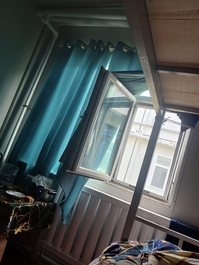 The image shows an interior view of a room, possibly a bedroom, with a window and curtains. A bunk bed frame is visible in the foreground. The window is open, allowing natural light to enter. Blue curtains are drawn to the side. On a table to the left, there is a plate with a piece of bread and a bowl, as well as some plastic bags. The overall impression is of a lived-in space, though no people are explicitly visible. The location is identified as Farab, Tajikistan.