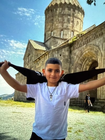 A young boy stands in front of a stone building, holding a black umbrella open above his head. He is wearing a white t-shirt with a cross necklace and dark shorts. The building appears to be an ancient monastery with a conical dome and stone arches. The boy is smiling and looking at the camera. The background shows a blue sky with some clouds and a mountainous landscape. The overall impression is of a sunny day at a historical site. This appears to be the Haghartsin Monastery, located in the Dilijan National Park of Armenia, not Artik.
