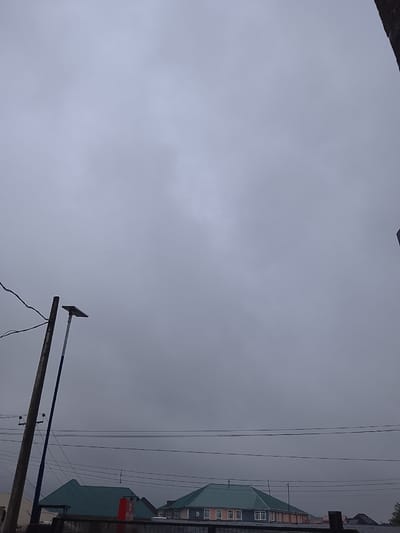 The image shows an overcast sky over a residential area in Rumuepirikom, Nigeria. In the foreground, a wooden utility pole stands to the left, with wires extending across the frame. Next to it, a modern lamppost with a solar panel is visible. Below the sky and utility poles, several houses with green roofs and visible windows are situated. A red water tank is positioned on top of one of the buildings. The overall atmosphere is dim, suggesting either early morning, late evening, or a cloudy day.