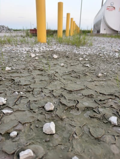 The image depicts a cracked mud surface with scattered white rocks and sparse green grass. In the background, several tall yellow cylindrical poles are visible, lined up in a row. Further in the distance, a large white cylindrical tank is present, suggesting an industrial or storage facility. The sky is a pale, overcast hue, indicating a diffused light condition. The ground appears dry and textured due to the cracking of the mud. No people are visible in the scene. The setting appears to be an outdoor industrial area in Cardwell, United States.
