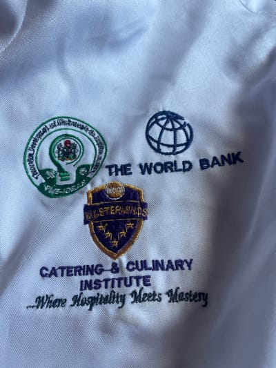 The image displays a close-up view of a white, textured fabric, likely a uniform or apron, adorned with multiple embroidered logos and text. In the upper left quadrant, a circular green emblem is embroidered, featuring the text "Fomota Development and Entrepreneurship" and "FME-IDEAS," with a smaller crest inside. To the upper right, a blue embroidered globe symbol is present, accompanied by "THE WORLD BANK" in blue capital letters below it. Centered slightly lower, a purple and gold shield-shaped emblem is embroidered, displaying "MASTERMINDS" across its upper section and containing stars on its lower portion. Below this shield, "CATERING & CULINARY INSTITUTE" is embroidered in purple capital letters, followed by the slogan "...Where Hospitality Meets Mastery" in a cursive, dark green/blue font. The fabric shows minor folds and a slightly coarse texture. The scene depicts embroidered insignia on a textile item in Mape, Nigeria.