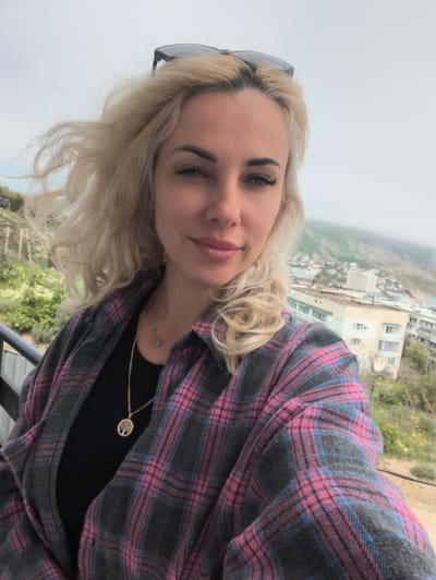A fair-skinned woman with blonde, shoulder-length hair is centered in the frame, posing for a selfie-style photo. She has sunglasses perched on her head and is wearing a black top and a grey and pink plaid flannel shirt. A gold necklace with a circular pendant is visible around her neck.

The woman is on a balcony, with a blurred landscape visible in the background. In the distance, a town or city is partially visible, with buildings and a hazy, overcast sky above. Vegetation is visible on the hills or slopes in the background. The photo is taken in the city of Орджоникидзевский поселковый совет, Ukraine.