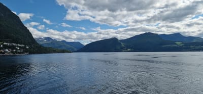 This is a wide-angle, horizontal landscape photograph taken from the water, likely a fjord, looking towards a mountainous coastline with a small village. The water in the foreground is calm with some ripples, reflecting the blue sky and scattered white clouds. On the left side, a steep, tree-covered mountain dominates the scene, with a cluster of white houses nestled along its base near the water's edge. Some of these houses have red roofs. In the background, a series of progressively distant, green-covered mountains rise, with some snow-capped peaks visible on the furthest ones. The sky is a vibrant blue with dramatic, puffy white clouds. The image appears to be taken during daylight hours. The location is likely in the vicinity of Volda, Norway, known for its fjords and mountainous terrain.