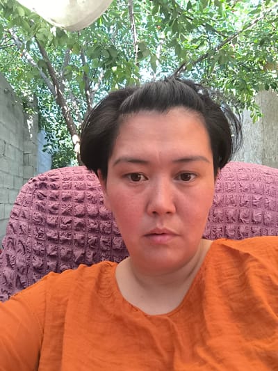 This is a selfie-style photo taken outdoors during the daytime. The subject is a person with dark hair and brown eyes, looking directly at the camera with a neutral expression. They are wearing an orange, short-sleeved shirt. The person is seated in a chair with a textured, purple cushion.

In the background, there is a tree with green leaves and some small red fruits, suggesting it might be a cherry tree. Behind the tree, a section of a grey, brick wall is visible, and to the right, another wall with a lighter, textured surface. A white, circular object, possibly a lampshade or a part of an umbrella, is partially visible at the top of the frame. The lighting suggests a sunny day, as indicated by the shadows and the brightness of the scene. The overall setting appears to be a backyard or garden area. The location context provided is Bereke, Kazakhstan. There is no visible text in the image.