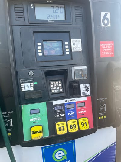 The image displays a close-up, slightly angled view of a black Gilbarco Veeder-Root fuel dispenser at a gas station in Midwest City, Oklahoma, United States. The upper digital display indicates "This Sale" with "$7.20" and "2.484 Gallons". A central screen within the pump housing shows a Coca-Cola advertisement featuring multiple bottles. Below this, a numeric keypad, a card slot reader, and a soiled contactless payment pad are visible. Adjacent to these are accessibility buttons with Braille, prompting the user to "SEE SCREEN ABOVE".
The lower section of the dispenser presents options for fuel types: "Clear Diesel", "Unlead" (87 octane, priced at "$2.899"), "Plus" (89 octane), and "Super" (91 octane), with a clear "NO ETHANOL" label above the gasoline selections. The pump is marked with a large white "6" and has "FJB" handwritten below it. A red "EMERGENCY SHUT OFF BUTTON IS LOCATED _ FT. OF FRONT DOOR" sign is affixed to the right side. A "PUMP CALIBRATION" sticker from the "OKLAHOMA CORPORATION COMMISSION" is partially visible. A green fuel hose extends from the left side, and a blue and white "e" logo (likely for the gas station chain) is visible at the bottom. The scene is illuminated by bright daylight. No individuals are present in the frame.