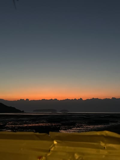This is a vertical, eye-level, medium shot of a sunset over a beach in Kertasari, Indonesia. The sky transitions from a dark navy blue at the top to a vibrant orange band just above the horizon, with the last vestiges of sunlight reflecting off the wet sand. The foreground is dominated by the blurry, out-of-focus yellow surface of an unidentified object, possibly a beach umbrella or a piece of furniture. In the midground, the exposed beach is dark and textured, showing ripples and patterns from receding tide. Islands are visible in the distance, silhouetted against the colorful sky. The overall mood is tranquil and serene, characteristic of a peaceful evening by the sea.