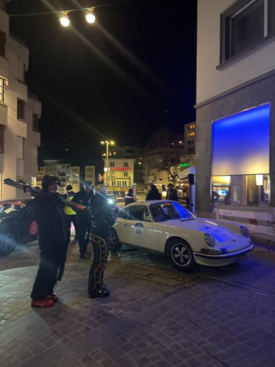 The image depicts a nighttime street scene in Sankt Moritz, Switzerland, likely during a winter event. In the immediate foreground, a person in dark winter attire, including red ski boots, stands facing right, carrying a pair of skis over their left shoulder. To their right, another individual is partially visible, wearing dark pants with gold star patterns and dark boots.

A vintage white Porsche 911 coupe, bearing the number "48" on its hood and driver's side door, occupies the mid-ground, positioned on a cobblestone street. The front of the car is illuminated by a prominent blue light emanating from a large vertical display on the adjacent building's facade. This building, on the right, features a light-colored upper section with a dark-framed window, and a darker base with the aforementioned blue-lit display.

To the left, another building with a light facade and multiple windows is visible. A dark-colored vehicle with a red "12" decal is partially visible behind the person carrying skis, parked on what appears to be a snow-covered patch of ground. Several individuals in the background, some wearing high-visibility vests, are clustered near this vehicle and further down the street.

Overhead, a string of warm white lights hangs diagonally across the upper left of the frame. The background reveals additional illuminated buildings, including one with decorative yellow string lights. The overall environment is an urban setting at night, characterized by artificial light sources and the presence of both event participants and organized personnel.