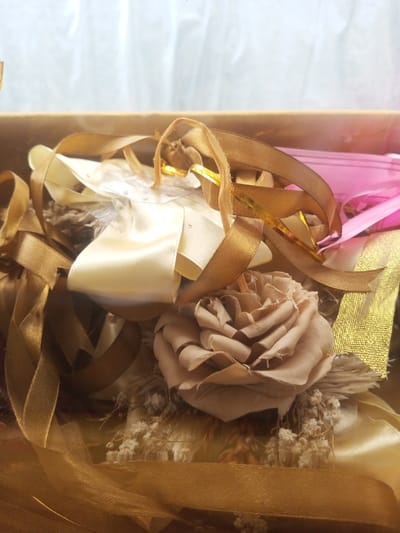 A close-up shot reveals a collection of craft materials nestled within a container. A neutral-toned rose lies prominently among golden and ivory ribbons, some sheer and others satin. Hints of pink and gold paper peek from beneath the layered materials. Dried baby's breath and feathery elements add texture. The overall effect is one of artistry and potential, suggesting a gift or a crafting project.