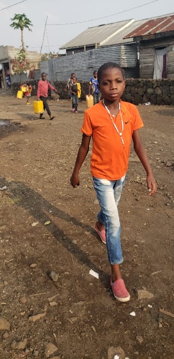 This image depicts a daytime scene in Buzove, Democratic Republic of the Congo, on an unpaved dirt road. The ground is rocky with scattered debris, including white fragments. In the foreground, a male child, approximately 8-12 years old, walks towards the viewer, slightly right of center. He wears an orange short-sleeved t-shirt with a black Nike logo on the left chest, light blue distressed jeans, and pink slip-on shoes. A white beaded necklace is visible around his neck. His gaze is directed forward. In the background, five other children are observed. To the left, a child in a red shirt walks away, carrying a yellow jerrycan in his left hand, with a smaller child behind him also carrying a yellow jerrycan. Further back, a child in a yellow jacket carries a yellow jerrycan. Another child in a blue shirt is partially visible behind them, also appearing to carry a jerrycan. The environment includes structures with corrugated metal roofs and walls of various materials, including metal sheeting and wood, and a low stone wall. A tall, slender tree with foliage concentrated at the top is visible on the left side of the frame.