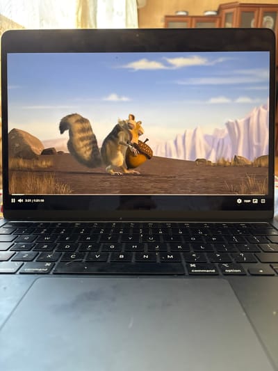 A laptop is positioned on a surface with a keyboard and trackpad visible. The laptop screen displays a scene from the animated movie "Ice Age," featuring the character Scrat holding an acorn. The background of the movie scene depicts a barren landscape with rocky formations and a pale blue sky with wispy clouds. The lower portion of the laptop screen shows playback controls, indicating the video is at 3:21 of 1:21:10 with a resolution of 720p. The overall environment suggests an indoor setting, possibly a home, with furniture visible in the background through a sheer curtain. There is no discernible location in Anton, Bulgaria associated with this image.