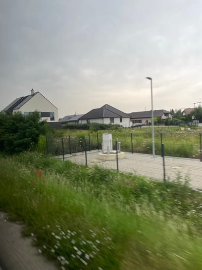 The image depicts a suburban residential area in Tyniec Mały, Poland. In the foreground, a fence runs horizontally across the frame, bordering a patch of overgrown grass and wildflowers. Beyond the fence, several modern houses are visible, characterized by their white facades, dark roofs, and large windows. One house, to the left, features a striking geometric design with contrasting black and white elements. A street lamp stands prominently in the mid-ground, accompanied by a white, box-like structure on a paved surface. The sky is overcast, suggesting a cloudy day. The overall impression is one of new development in a semi-rural setting, with a focus on contemporary architectural styles.