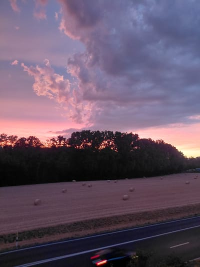 A rural landscape is depicted during sunset. The sky is a vibrant mix of pink, purple, and gray clouds. In the foreground, a field with several hay bales is visible. A line of trees runs across the middle ground, separating the field from the sky. A highway is in the lower right corner of the image, with a vehicle's taillights blurred due to motion. The scene appears to be in the vicinity of Ecublens, Switzerland, judging by the hay bales in the fields and the typical rural scenery.