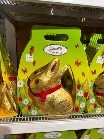 The image displays a retail store shelf containing multiple large Lindt "Goldhase" (Golden Hare) chocolate bunnies, presented in their original packaging. Each chocolate bunny is wrapped in reflective gold foil and features a red ribbon with a small bell around its neck. These are housed within individual bright green cardboard boxes, designed with an integrated handle cutout at the top. The boxes are decorated with illustrations of white daisies, red and yellow tulips, and red butterflies, implying a spring or Easter theme. A prominent oval label on each box reads "Lindt GOLDHASE" in black text. A white price tag is affixed to the upper left portion of the green packaging, displaying "Fr. 39.95" and a unit price of "(100g = 7.99)", indicating Swiss Francs and confirming the location as Switzerland. The products are arranged on a white, open-grid metal retail shelf. Another identical shelf is visible above, holding more products, partially out of frame. The background consists of brown wooden paneling. The specific location within the city of Fribourg - Freiburg, Switzerland, cannot be determined from the visual evidence beyond the country and general retail environment. No individuals are present in the scene.