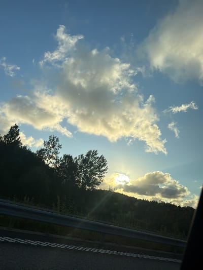 The image shows a view from a car, looking out towards a hillside covered in trees. The sun is low in the sky, partially obscured by clouds, casting a warm glow. There is a guardrail visible in the foreground, separating the road from the hillside. The road itself has double white dashed lines, indicating a lane separation. The sky is blue with scattered, fluffy clouds. The overall scene suggests a scenic drive, possibly during sunset, in or near Ödsmål, Sweden, given the natural landscape and vegetation.