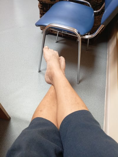 A first-person perspective shot shows a person's legs crossed at the ankles, resting on a speckled gray floor. They are wearing dark blue shorts. In the background, a blue chair with chrome legs is partially visible, as is the leg of another chair and a patterned fabric couch. The environment appears to be indoors, possibly a waiting room or public space. The location is not specified in the image or provided context.