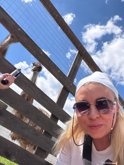 This low-angle selfie, taken in Rassvet, Russia, captures a close-up of a blonde woman interacting with an emu in an outdoor enclosure. The image is tilted, with the woman's face positioned in the upper right quadrant, looking down towards the viewer and the emu. She wears a white bandana with a subtle, unreadable pattern, large dark sunglasses connected by a beaded chain with small heart-shaped accents, and a white top. Her blonde hair flows around her face, and she has a gentle smile.
In the lower center of the frame, her right hand is visible, holding a black monopod or selfie stick, suggesting she is taking the picture herself. A sturdy wooden fence with wire mesh diagonally crosses the frame, acting as a barrier between the woman and the animal. Through an opening in this fence, the head and part of the neck of a large emu are clearly visible in the mid-left portion of the image. The emu has dark, shaggy feathers on its head and appears to be looking upwards.
The background consists of a bright blue sky dotted with scattered white, fluffy clouds, indicating a clear and sunny day. Parts of the emu's brown, feathery body are also discernible through other sections of the wooden fence. The scene depicts a casual, interactive moment at what appears to be an animal park or farm.