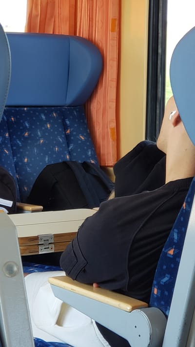The image shows the interior of what appears to be a bus or train carriage. The scene is dominated by blue seats with an orange and blue patterned fabric. In the foreground, a person is partially visible, wearing dark clothing. Their head is tilted down, and their face is not clearly visible. A light-colored wooden tray table is extended, and the person's hand rests on it. To the left, a blue seatback is prominent, with orange curtains visible behind it. The overall lighting suggests daytime. The location is identified as Brunovce, Slovakia.