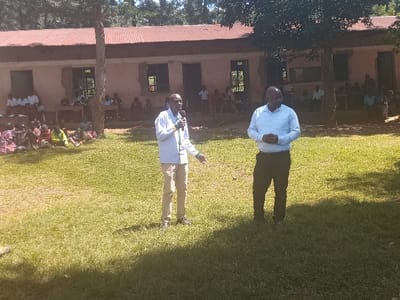 The image depicts an outdoor gathering in the Idakho South ward, Kenya, on a sunny day. In the foreground, two adult males stand on a field of green grass. The man on the left, wearing a light blue long-sleeved shirt and tan trousers, holds a black microphone in his right hand and gestures with his left. The man on the right, dressed in a light blue long-sleeved shirt and dark trousers, stands with his hands clasped in front of him. Behind them, a single-story building with a reddish-brown corrugated metal roof and light-colored walls features multiple rectangular window openings and doorways. Approximately 15-20 individuals, primarily children in various attire, including some in school uniforms, are seated on the grass to the left of the scene, facing the two men. Additional individuals, including adults, are visible seated within the building's openings to the right. A large tree with dark green foliage partially obscures the building between the two standing men and the background group. The ground is covered in green grass, with natural sunlight casting shadows.