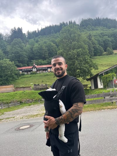 This image depicts a man standing outdoors in a scenic, hilly landscape. The man, who appears to be of adult age, is smiling and looking towards the camera. He has numerous tattoos on his left arm and is wearing a black t-shirt and black pants, with a black backpack or bag slung over his shoulder. He is holding a small object in his right hand, which resembles a spray can.
In his arms, the man is holding a baby, whose feet and legs are visible. The baby is wearing white socks with a pattern on them. The man's posture and smile suggest a sense of pride and affection.
The background reveals a lush green environment with rolling hills covered in dense trees, indicative of a forest. A traditional-looking building, possibly a house or a guesthouse with a red roof, is situated on the hillside, surrounded by greenery. There is a paved pathway or road in the foreground where the man is standing, with a manhole cover visible on the ground. A wooden fence runs along the side of the pathway.
The sky is overcast, suggesting a cloudy or potentially rainy day. The overall impression is one of a serene, natural setting, possibly in a rural area of Germany, given the location context of Ettal. The man's engagement with the baby and the peaceful backdrop create a heartwarming and intimate scene.