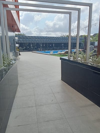 The image shows a view through a white frame structure, looking out to an outdoor area. The frame is constructed from three white rectangular beams, forming a rectangular shape. The frame is positioned between a black wall to the left and a black wall with a dark grey tiled surface to the right. The outdoor area is visible through the frame. The tiled surface is light grey, with a smooth finish. The outdoor area has a blue swimming pool in the center. The pool has a black tiled border and is surrounded by a grassed area. There are three black rectangular sunbeds on the grass to the right of the pool. A row of dark grey rectangular structures are seen in the background, likely covered by some form of fabric. The image captures a section of the surrounding environment in Ujevwu, Nigeria, with the coordinates of 5.472 latitude and 5.817 longitude. One person is visible in the background to the right, standing close to a black structure, partially obscured by a large potted plant. The person appears to be wearing a white shirt and has dark hair. There is a string of white lights running horizontally across the top of the dark grey structures. The sky is visible in the background, with a few clouds in the sky.