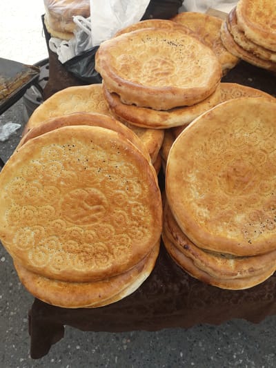An overhead shot displays a large quantity of round, golden-brown flatbreads, characteristic of Central Asian "non" or "naan," arranged outdoors in Ghafurov, Tajikistan. The bread is stacked in several piles, filling most of the frame. Many loaves feature intricate, pressed patterns in their centers, often with circular or floral motifs and a raised central dome, while their edges are thicker and slightly raised. Some of the bread is sprinkled with black nigella seeds or white sesame seeds.

The display rests partially on a dark brown fabric or rug. A grey, speckled ground, likely asphalt or concrete, is visible on the right and bottom edges of the frame. Partially visible white and black plastic bags in the lower left suggest other market goods or packaging. The even lighting indicates daytime. The scene depicts a typical market or roadside stand where freshly baked bread is offered for sale. No people or visible text are present in the image.
