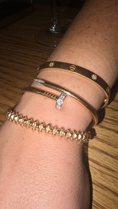In Trikomo, Northern Cyprus, a close-up, eye-level shot shows a person's forearm adorned with three gold bracelets. The uppermost bracelet is a thin gold band with inset diamonds and the Cartier "O" logo repeated. Beneath it is a thicker gold band, also with the Cartier "O" logo and inset diamonds. The lowest bracelet is a thick, gold chain composed of interlocking spiked links. The forearm is lightly tanned and shows faint freckles. The bracelets are positioned on the forearm as it rests on a textured wooden surface, likely a table. A blurred background reveals the stem of a wine glass.