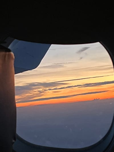 The image shows a view through an airplane window. The sky is visible, exhibiting colors of orange, white, and dark blue, suggesting either sunrise or sunset. Part of the airplane's wing is visible in the lower left corner of the window. The view does not seem to be showing any feature associated with the city of Ernzen, Luxembourg. The image provides no information about people, subjects, interactions or objects.