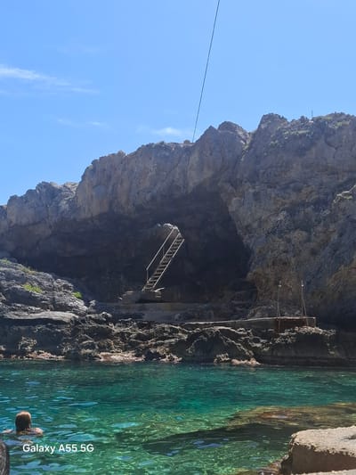 The image depicts a grotto near Plakias, Greece. A tall rock formation, mainly gray and covered in rocky outcroppings, faces the camera, concealing a dark cave underneath. A metal ladder is installed inside the cave. To the right of the rock formation, clear green water extends across the frame. In the water, a person's head can be seen. The sky is blue with a few wisps of clouds. A thin wire stretches across the upper left of the frame. A "Galaxy A55 5G" watermark is visible in the bottom right corner.