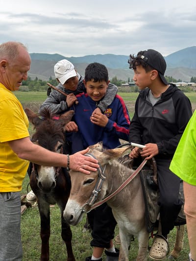 In Shabdan, Kyrgyzstan, on an overcast day, a group of individuals, including three young males and an adult male, interact with two donkeys. The adult male, wearing a yellow t-shirt and grey pants, is reaching out to touch the head of a dark brown donkey. One of the young males, wearing a white baseball cap and a grey hooded jacket, is seated on the brown donkey, while another young male, wearing a blue jacket with red trim, is perched on his shoulders. The third young male, wearing a black baseball cap and a black Nike jacket, is seated on a lighter brown donkey to the right. Both donkeys are equipped with saddles and bridles. The background features rolling hills and a few buildings, suggesting a rural or semi-rural setting.