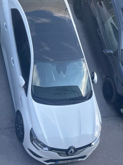 The scene depicts an overhead view of a white Renault passenger vehicle parked on an asphalt surface in Logatec, Slovenia. The white vehicle features a black panoramic roof and multi-spoke dark alloy wheels. Its front license plate, "LJ 15-BBJ", is visible. To the immediate right of the white vehicle, a dark-colored, possibly black, sedan or hatchback is partially visible, also parked on the asphalt, displaying black multi-spoke rims. Reflections of a cloudy sky and architectural elements are present on the vehicles' windows and body panels. No individuals are present, and the vehicles appear stationary.