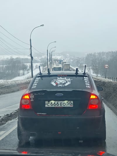 The image depicts the rear of a dark-colored Ford Focus sedan on a wet, snow-lined road during an overcast winter day in Podosinki, Russia. The vehicle is stationary or moving slowly, indicated by its illuminated red brake lights. Its Russian license plate is displayed as 'C 666 EO 50 RUS'.
The rear window of the Ford Focus features multiple white stickers: a large graphic of a crowned lion's head with red accents in the center; an Armenian cross and the text "ԽԱՂԱՂՈՒԹՅՈՒՆ ԵՎ ՓՐԿՈՒԹՅՈՒՆ" (Armenian for "Peace and Salvation") vertically on the left; and the vertical text "解救和保佑" (Chinese for "Deliverance and Blessing") on the right. Below the Ford badge, a smaller white sticker reads "Kasumi. Ты мне очень нужен" (Russian for "Kasumi. I really need you"). Four black vertical posts, consistent with antenna mounts or a roof rack system, protrude from the vehicle's roof. A sticker on the lower left of the rear bumper reads "ЗАТО НЕ В КРЕДИТ" (Russian for "BUT NOT ON CREDIT"). Another small, illegible white sticker is present on the lower right of the bumper.
In the background, a line of semi-trailer trucks and other vehicles extends down the road, suggesting traffic congestion. The road is bordered by a concrete barrier on the right and power lines on the left, with a series of streetlights extending into the distance. Snow covers the ground and the leafless trees on the adjacent hillsides. A circular road sign with a red border and white background, containing two horizontal red bars, is visible on the right side of the road.
