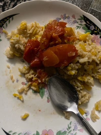 The image displays a close-up view of a meal served on a white, floral-patterned plate. The plate's rim features an ornate design with pink, purple, and green flowers, and black leaf accents. The main dish consists of a generous portion of rice mixed with small, yellow, cubed vegetables, possibly squash or bell peppers. On top of and alongside the rice are several pieces of stewed or roasted red tomatoes, some appearing whole or halved, while others are broken down, with visible slivers of white onion. A silver metal spoon rests on the right side of the plate, its bowl partially submerged in the food. The background immediately surrounding the plate is a textured, possibly gray or metallic surface, suggesting an indoor setting. The scene depicts a meal, potentially in progress or recently served, in Yangikishlak, Uzbekistan. No people or visible text are present in the image.