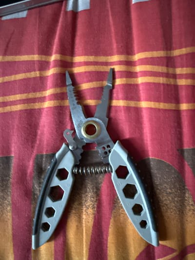 An overhead shot shows a multi-tool pliers lying open on a red and orange striped fabric. The fabric appears to be a towel or blanket, with a slight sheen and a textured weave. The multi-tool is made of silver-colored metal with gray plastic grips. The pliers are in the center of the frame, with their handles spread apart. The jaws of the pliers have a serrated edge and a small cutting edge. Between the jaws is a circular opening with a gold-colored ring. The handles of the pliers have several hexagonal cutouts of varying sizes, labeled with numbers such as 10, 13, 9, 11, and 12. A coiled spring connects the two halves of the tool, allowing the jaws to spring open. The overall scene is well-lit, with no shadows indicating any specific time of day. There are no people or other objects visible in the image. The image is focused on the multi-tool, highlighting its details and design. The location is identified as Морозовка, Russia, but the image itself provides no visual cues to support this specific location.