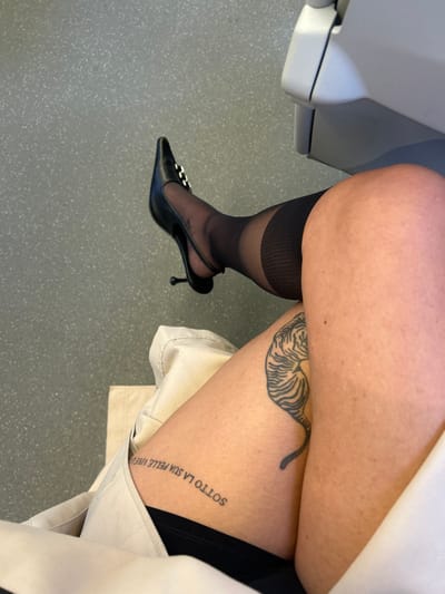 The image presents a close-up, high-angle perspective of an individual's lower body, observed in St Albans, United Kingdom. The left leg, extended towards the upper left of the frame, is fitted with a black, pointed-toe stiletto heel featuring a silver-toned, rectangular buckle on the vamp and a slender, flared black heel. This leg is covered by sheer black hosiery, which exhibits a distinct, thicker, opaque black band encircling the upper thigh. The right leg, partially visible, displays bare skin on the thigh. A black-line tattoo of a crouching tiger is visible on the outer aspect of the right thigh, with its head directed upwards. An additional tattoo, text reading "SOTTO LA SUA PELLE VIVE...", is situated upside down on the inner-upper right thigh, positioned above a dark fabric undergarment. A light beige or off-white garment is draped across the lap and lower right thigh. The background floor is a uniform light grey with subtle speckling, and a segment of a white or very light grey, curved object, likely part of a seat or piece of equipment, is present in the upper right corner.