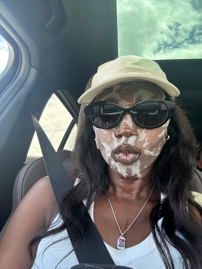 A person wearing a face mask and sunglasses is in a car. The person is wearing a light-colored hat, a white top, and a silver necklace with a purple pendant. The seatbelt is fastened. The sky is visible through the car's sunroof. The location is in a car, likely somewhere in Thousand Palms, United States of America.