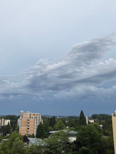 An overcast sky looms over a residential area in Sindelfingen, Germany. In the mid-ground, a multi-story apartment building with a beige and orange facade stands amidst dense green trees. Other buildings and structures are visible in the background, partially obscured by foliage and the cloudy sky. The foreground is dominated by the tops of numerous trees. The overall scene depicts a cloudy day in a suburban environment.